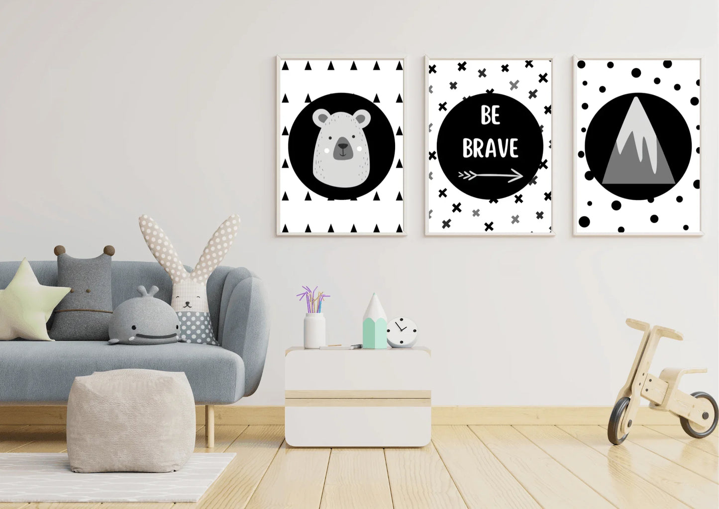 Three framed monochrome nursery prints, depicting a bear, "Be Brave" text, and a mountain, hanging on a white wall above a small side table and next to a blue couch and children's toys.