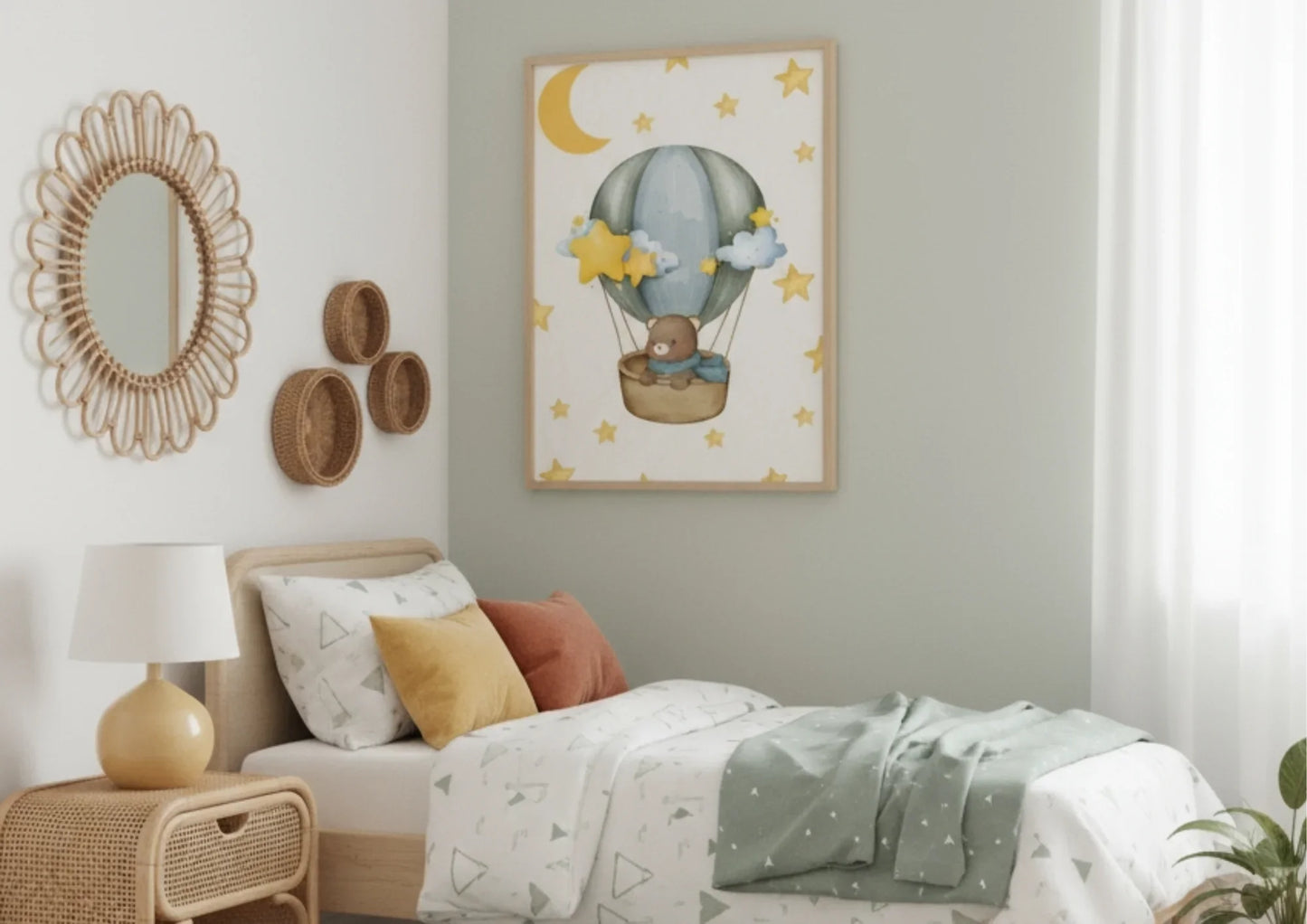 A vertical framed watercolor print hanging above a bed with green and white patterned bedding. The artwork features a brown teddy bear wearing a blue scarf, sitting in a wicker basket carried by a gray and blue hot air balloon. The light background is patterned with yellow stars and a large crescent moon. The room has walls painted in white and a muted sage green, and is decorated in a bohemian style with a round rattan mirror, rattan nightstand, and three small woven wall baskets.