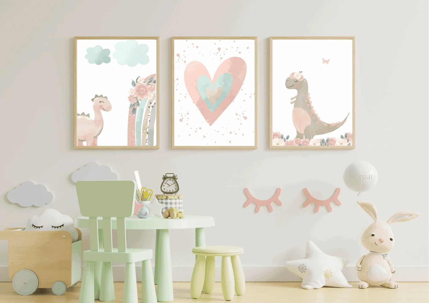 A trio of framed art prints hangs on a white wall above a small, light green table and chair set. The prints feature a whimsical, watercolor-style set with a pink dinosaur, a large pink and teal heart, and another dinosaur with a floral crown. The room is decorated with a cloud-shaped toy bin, a star-shaped pillow, and a stuffed bunny with a balloon.