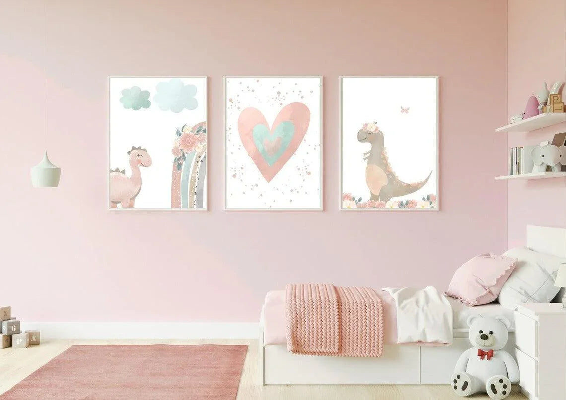 A set of three framed art prints hangs on a light pink wall in a child's bedroom. The prints feature a watercolor-style illustration of a pink dinosaur, a large pink and teal heart, and a gray dinosaur with a floral crown, all in a whimsical, boho style.