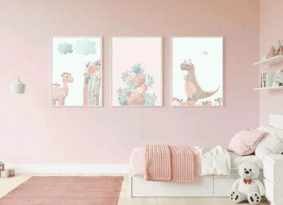 Three framed pastel dinosaur and cactus art prints on a light pink wall above a child's bed with a knitted blanket in a pink bedroom.