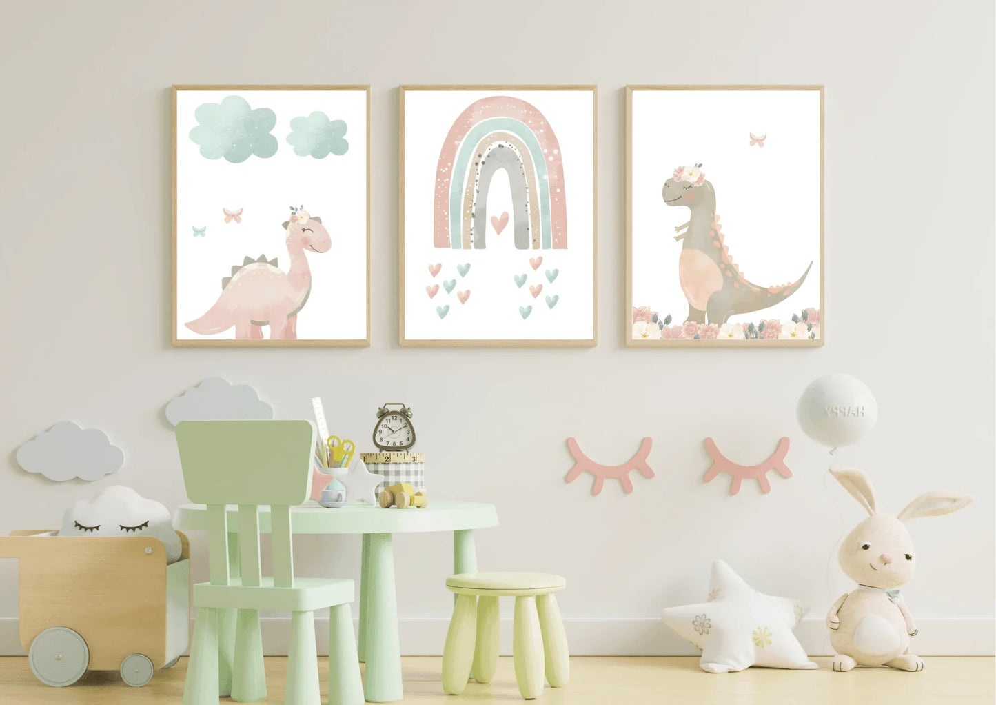 A three-panel set of digital nursery art prints featuring a pastel pink dinosaur with a crown, a soft rainbow with hearts, and a friendly dinosaur surrounded by flowers. The prints are framed and hang above a small green table and chair set in a child's room.