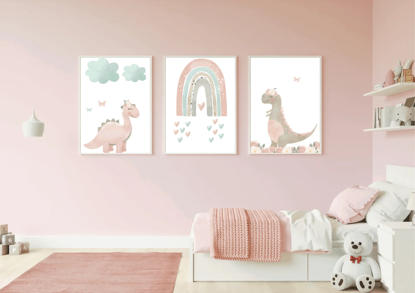 A child's bedroom with light pink walls and a matching pink rug. A set of three framed digital art prints hang above a small white bed with a pink blanket. The art features pastel pink and brown dinosaurs and a rainbow. A white teddy bear sits on the floor next to the bed.