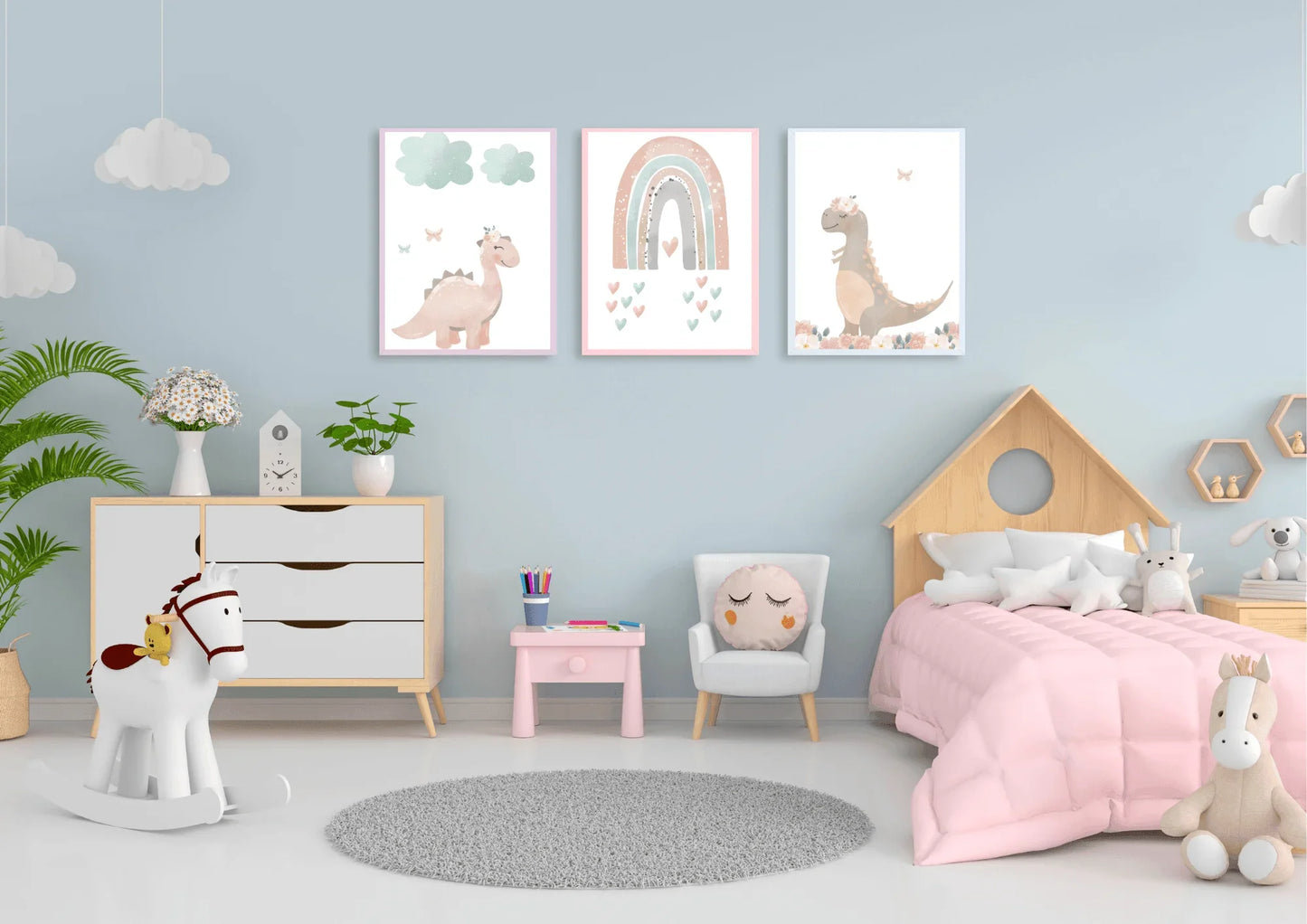 A set of three framed nursery art prints depicting pastel dinosaurs and a rainbow, hanging on a light blue wall above a dresser and a child's bed. The room is decorated in a soft color palette with a rocking horse, a small table and chair, and a fluffy rug.