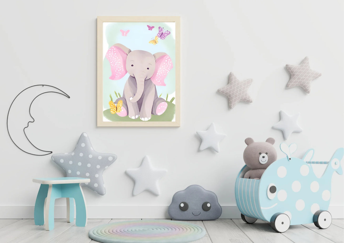 A framed digital art print of a baby elephant with pink ears and butterflies, hanging on a white wall in a nursery. A crescent moon wire decoration is on the left, and a toy box shaped like a blue whale with white spots is on the right. Star-shaped pillows are on the floor and wall.