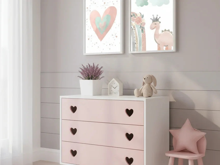 A close-up of a modern nursery with a white dresser that has pink drawers and heart-shaped handles. On the wall above, two framed prints show a watercolor heart and a cute pink dinosaur with a crown, adding a charming and playful touch to the room.