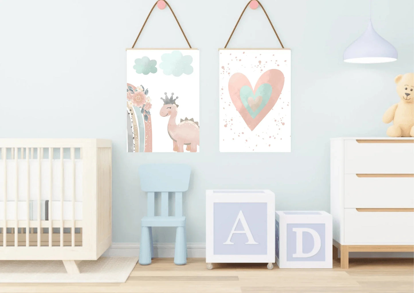A playful baby's room with a light blue wall. Two framed prints hang from the wall with rope, showing a cute pink dinosaur with a crown and a watercolor heart. The room also features a crib, a small blue chair, and large letter blocks, creating a sweet and inviting space.