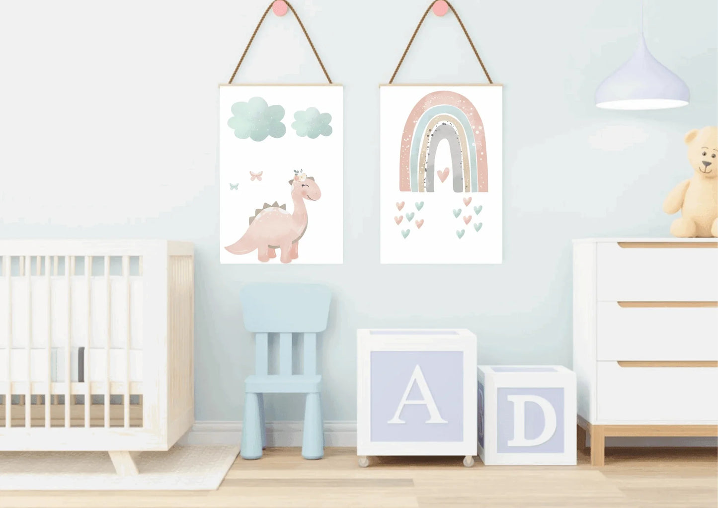 Two hanging pastel prints (pink dinosaur with clouds, and a rainbow with hearts) on a white and blue wall above a baby crib and dresser.