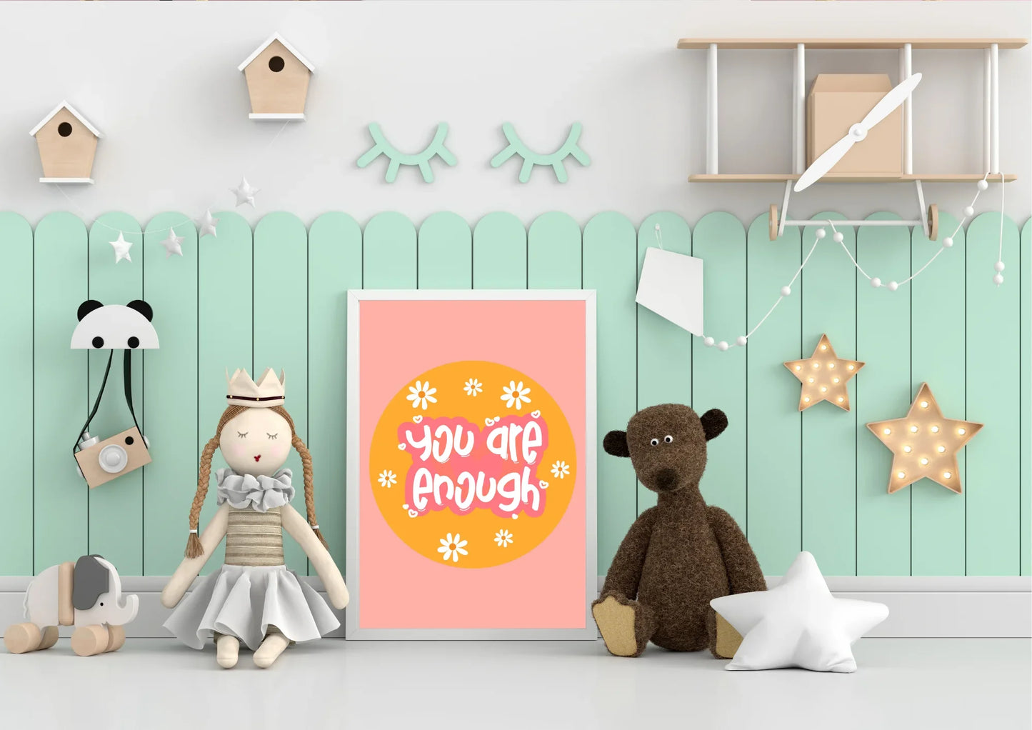 A framed "You are enough" art print on a two-toned wall in a child's playroom with a teddy bear and star lights.