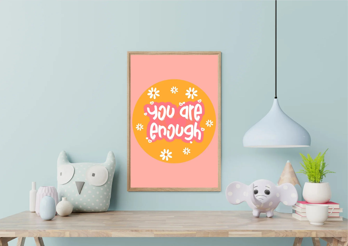 A wooden-framed "You are enough" art print on a light blue wall in a child's room with an owl and elephant pillow.