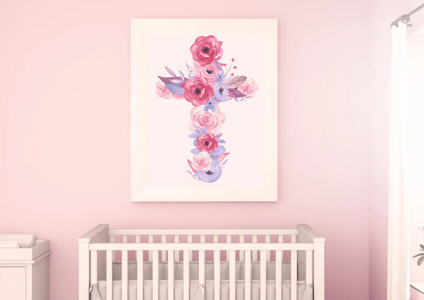 A close-up view of a nursery with a light pink wall. A white framed art print of a cross made of pink and purple watercolor flowers hangs above a white crib. A white dresser is partially visible on the left.