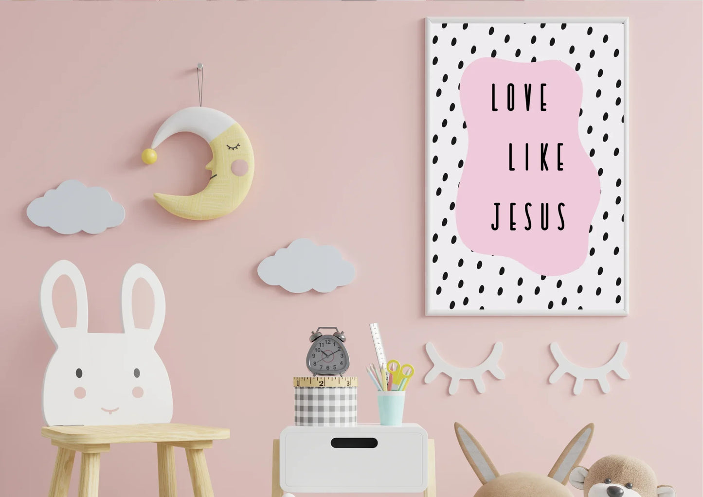 A framed "Love like Jesus" art print on a pink wall in a child's nursery with a sleepy moon and clouds.