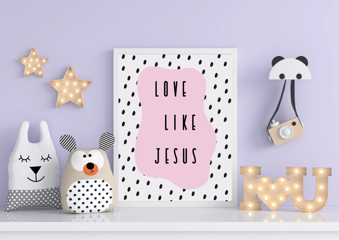A framed "Love like Jesus" art print on a shelf with pillows and light-up stars on a light purple wall.