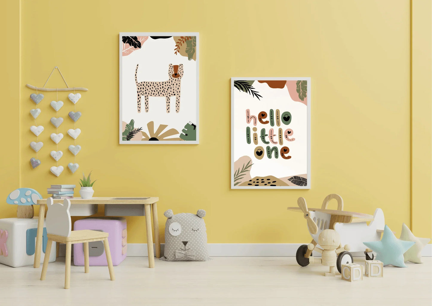 A bright nursery with yellow walls, featuring a two-piece framed art set with a boho jungle theme, including a cheetah/leopard print and "hello little one." The room has a small wooden table and chair set, a heart wall hanging, and wooden toy airplane.