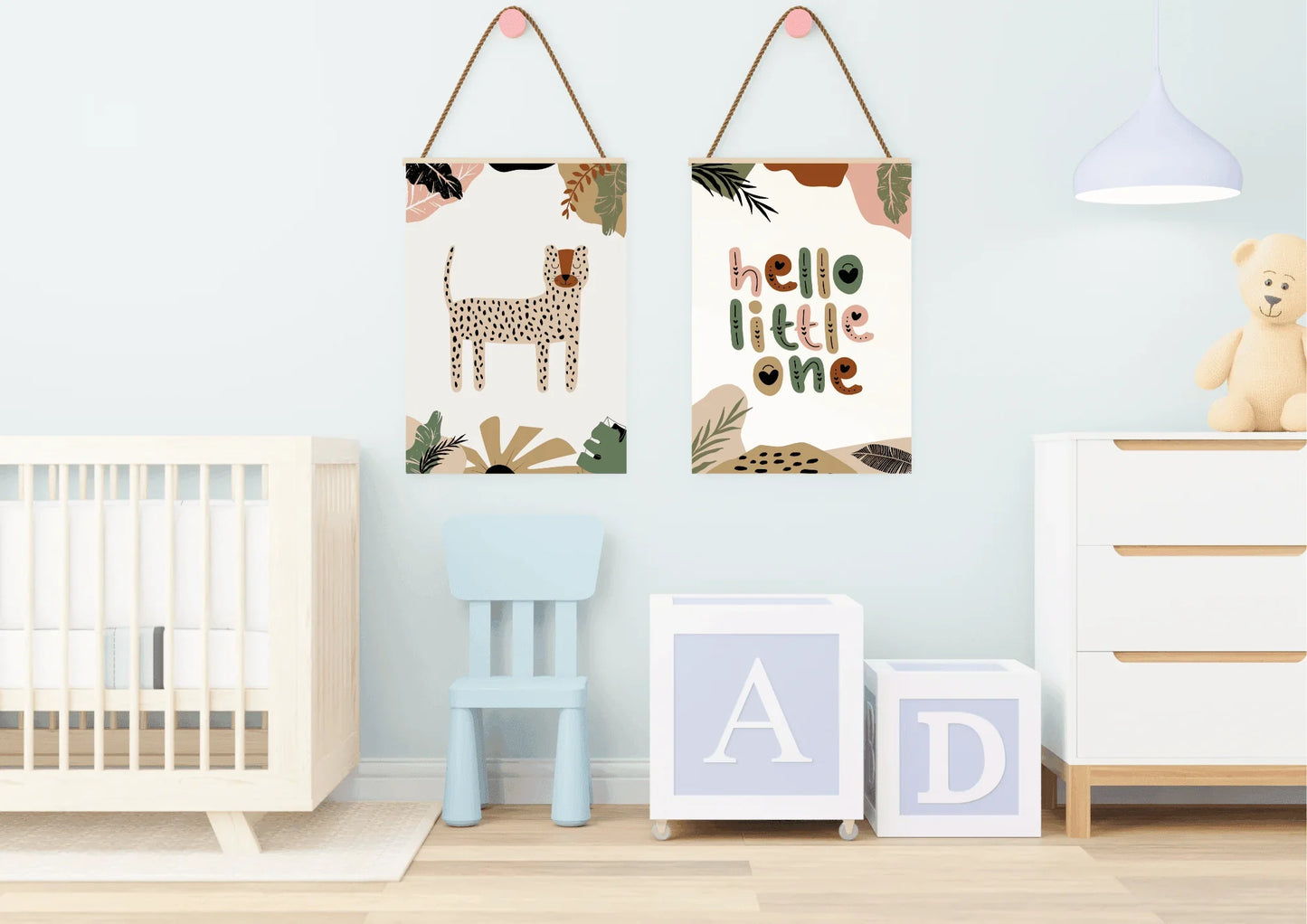 A nursery with light blue walls, featuring a two-piece framed art set with a boho jungle theme hung from pink ribbons. The room contains a light wooden crib, a small blue chair, and white toy blocks with letters A and D.