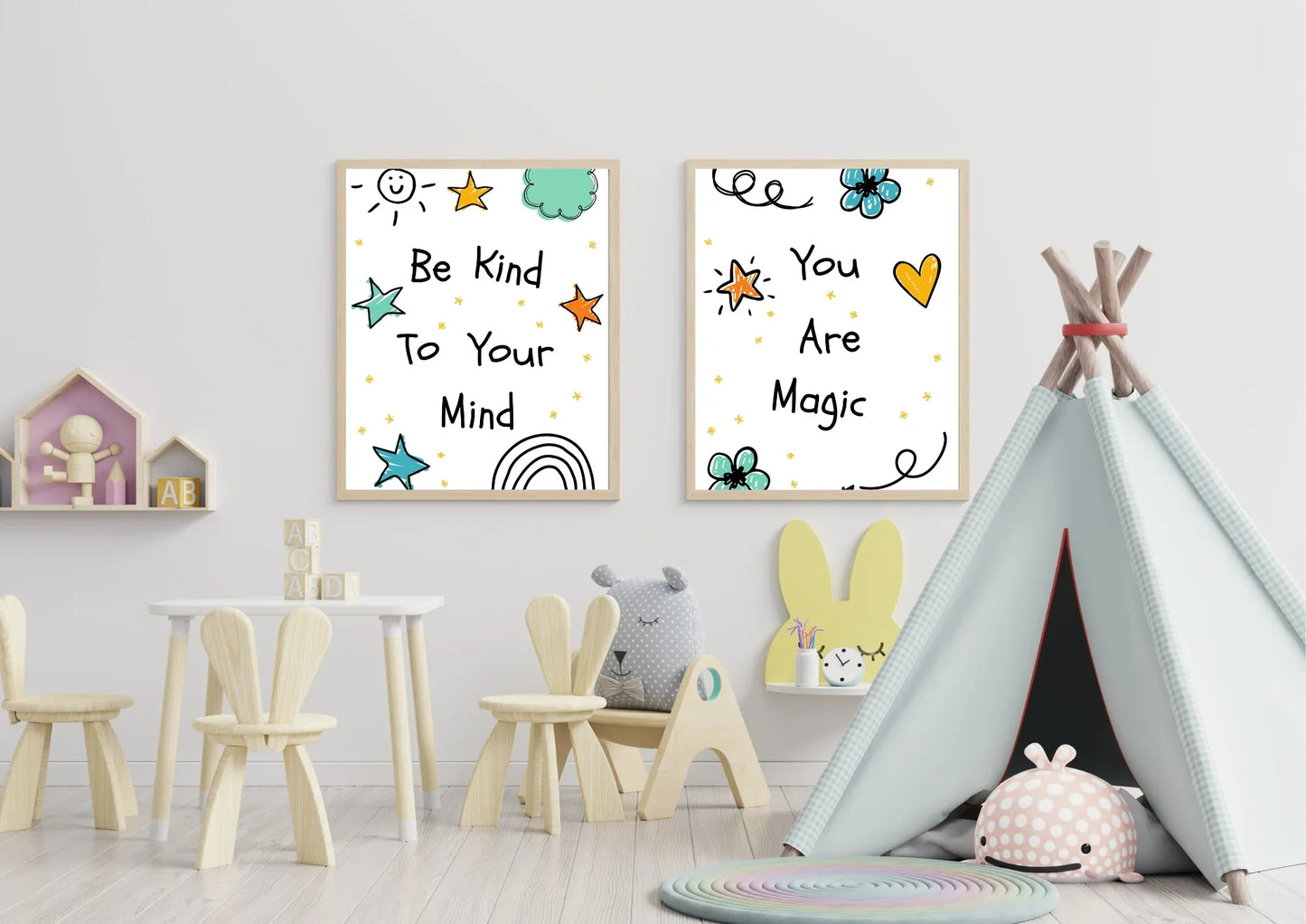 A two-panel set of framed digital art prints with motivational quotes, hanging on a white wall in a child's playroom. The left print says "Be Kind To Your Mind" and the right one says "You Are Magic." A small wooden table and chairs, a teepee, and various toys are in the room.