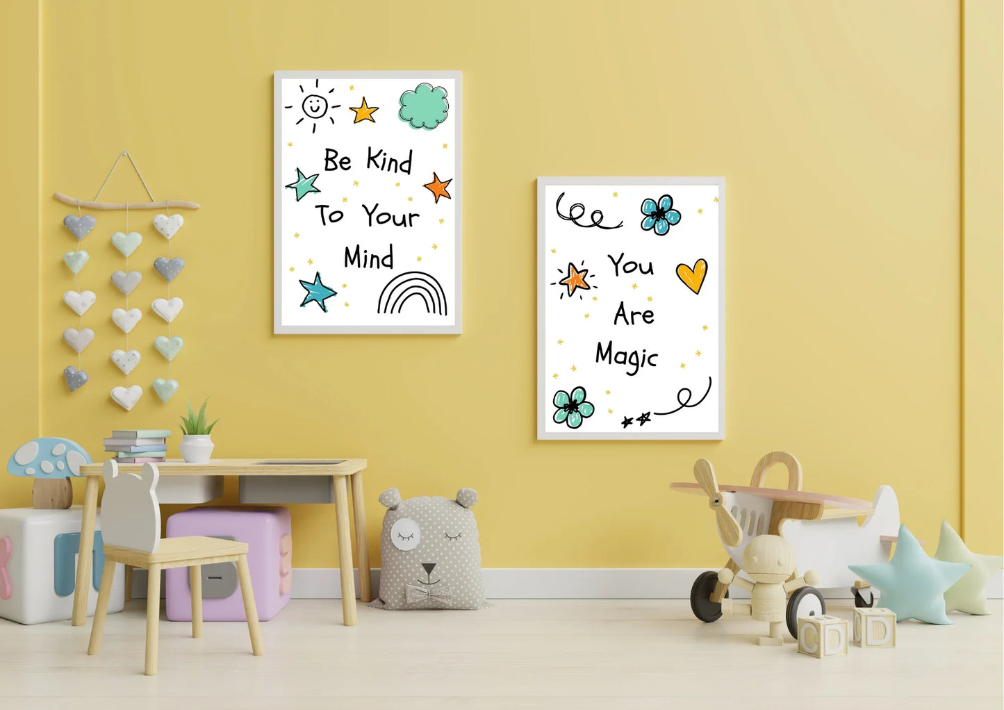 A pair of framed digital art prints with whimsical doodles and positive affirmations, hanging on a yellow wall. The left print says "Be Kind To Your Mind" and the right one says "You Are Magic." A small wooden desk with a chair and a toy airplane are in the room.