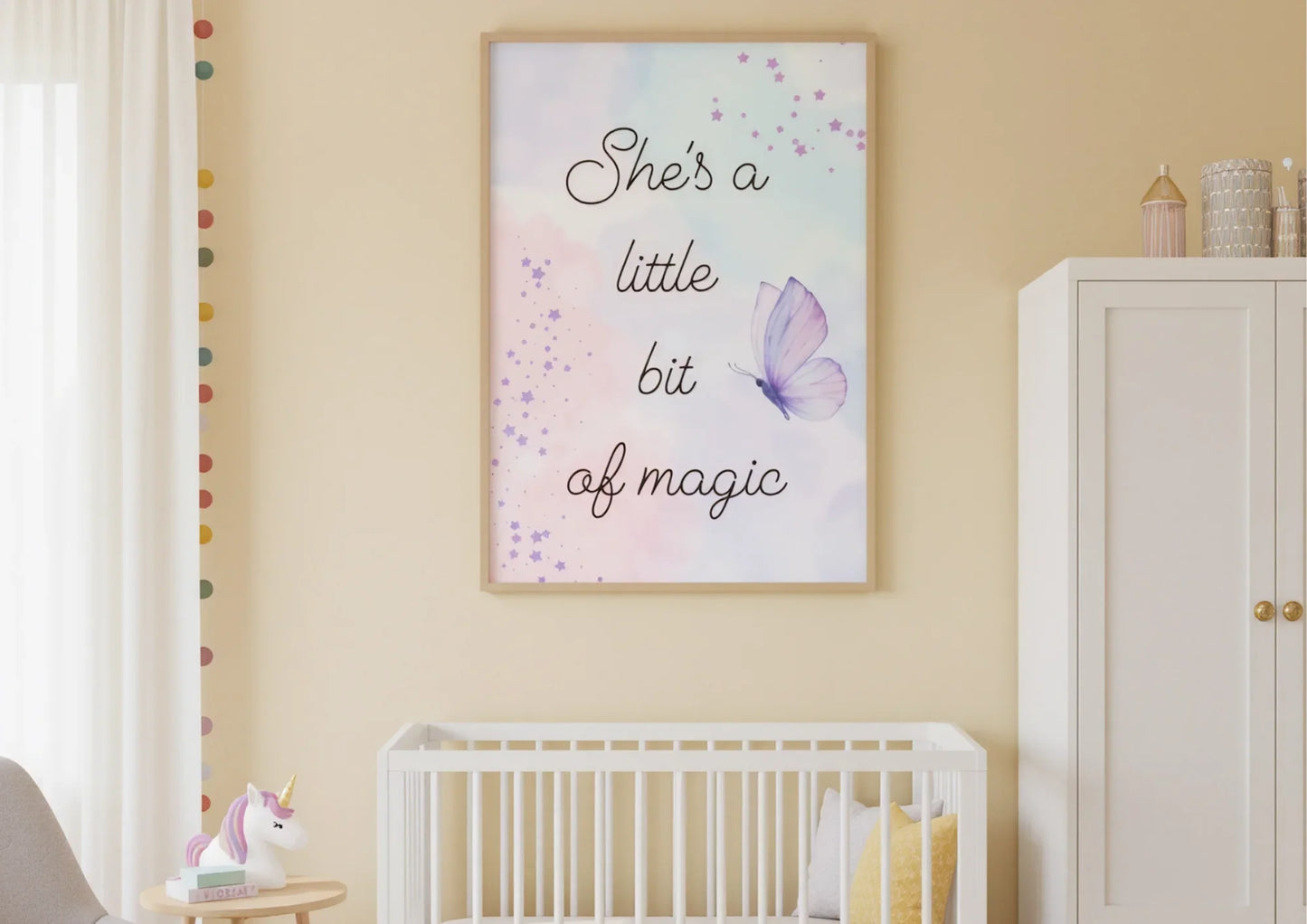 A charming nursery with soft yellow walls. A framed art print with a watercolor background and the words "She's a little bit of magic" is hanging above a white crib with a light yellow pillow. A white wardrobe with gold handles is on the right, and a unicorn lamp is on a small table on the left.
