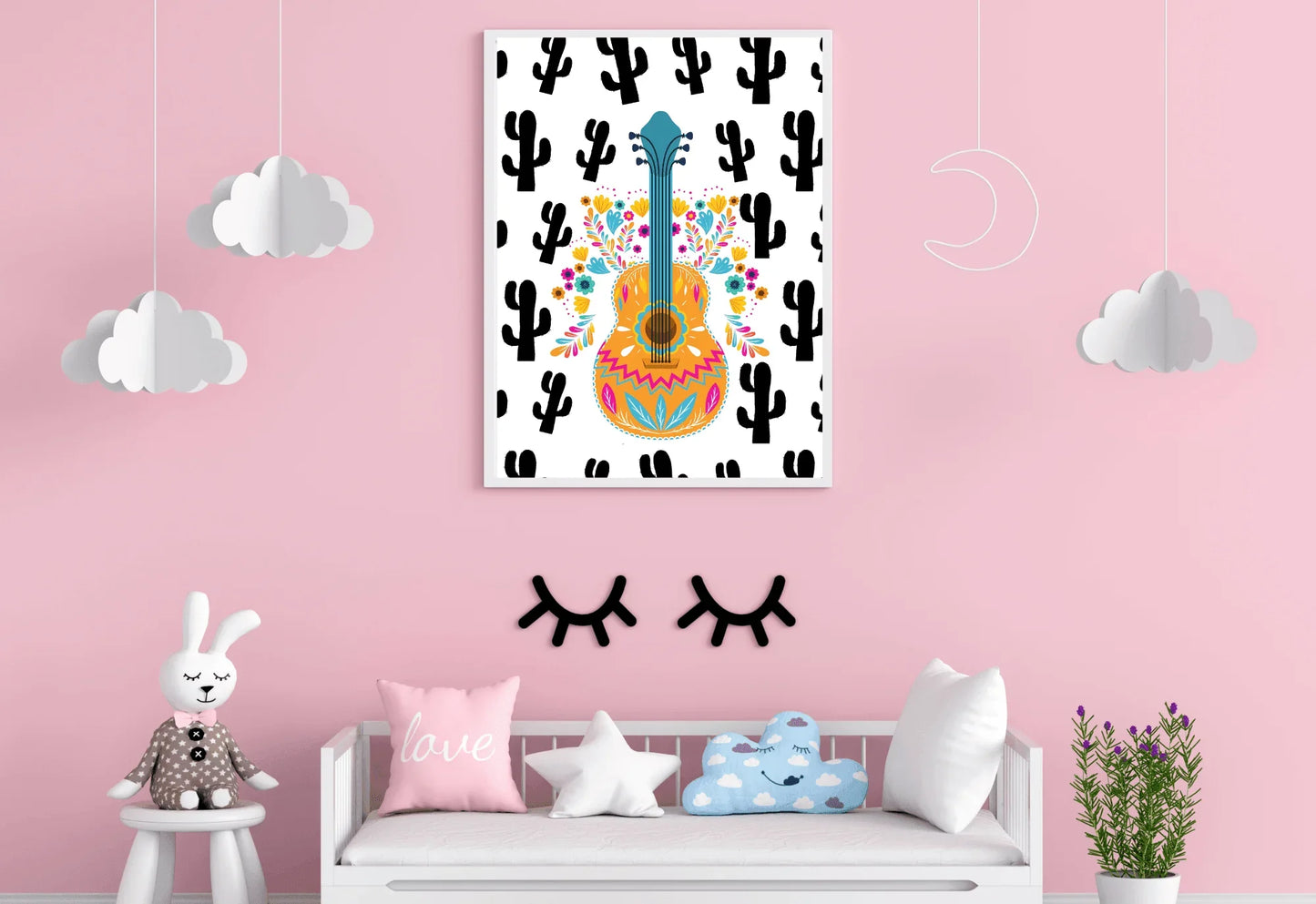 A framed boho folk art guitar print with a black cactus background, hanging on a light pink wall above a white bench with pillows.