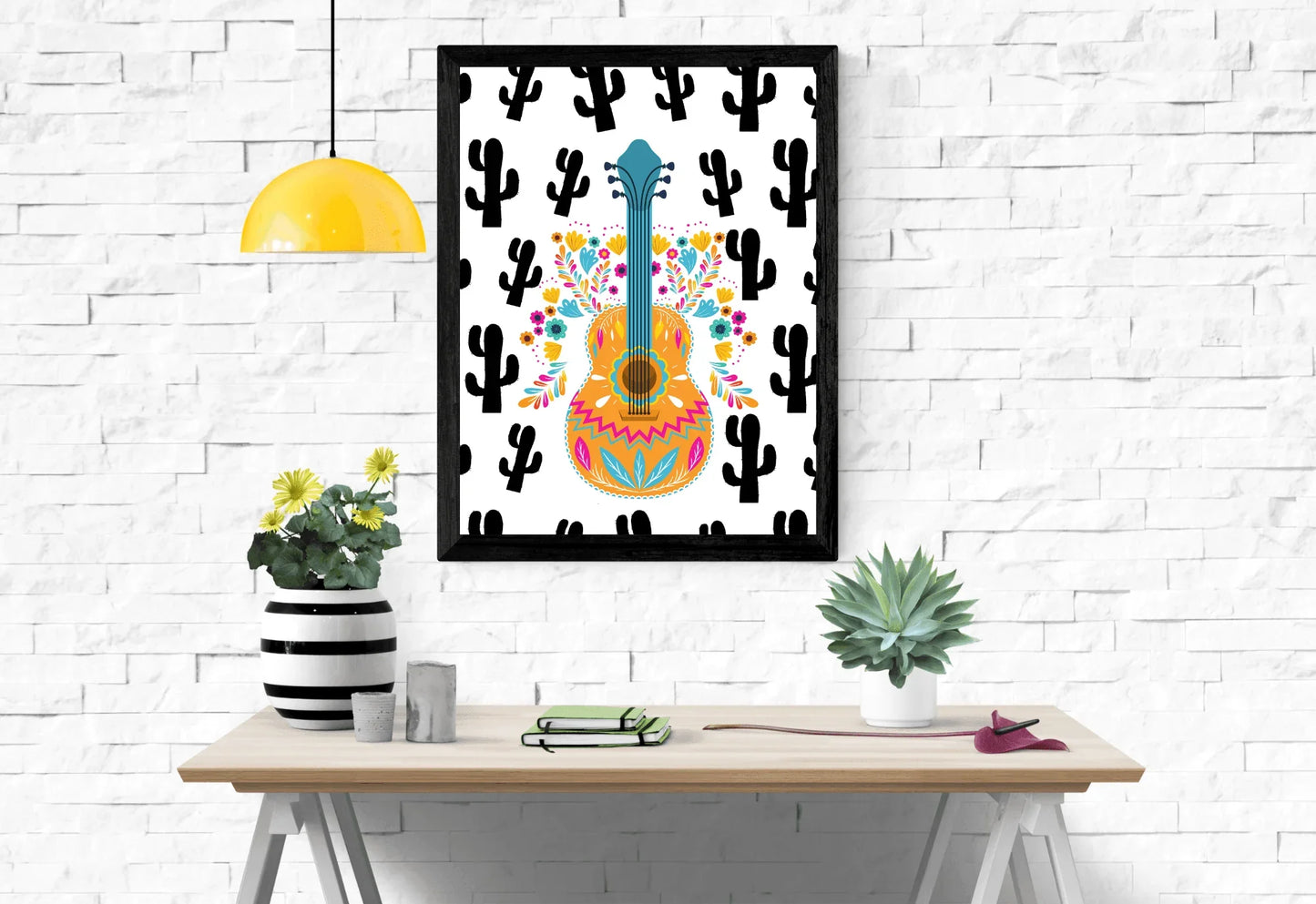A framed boho folk art guitar print with a black cactus background, hanging on a white brick wall above a wooden desk with plants.