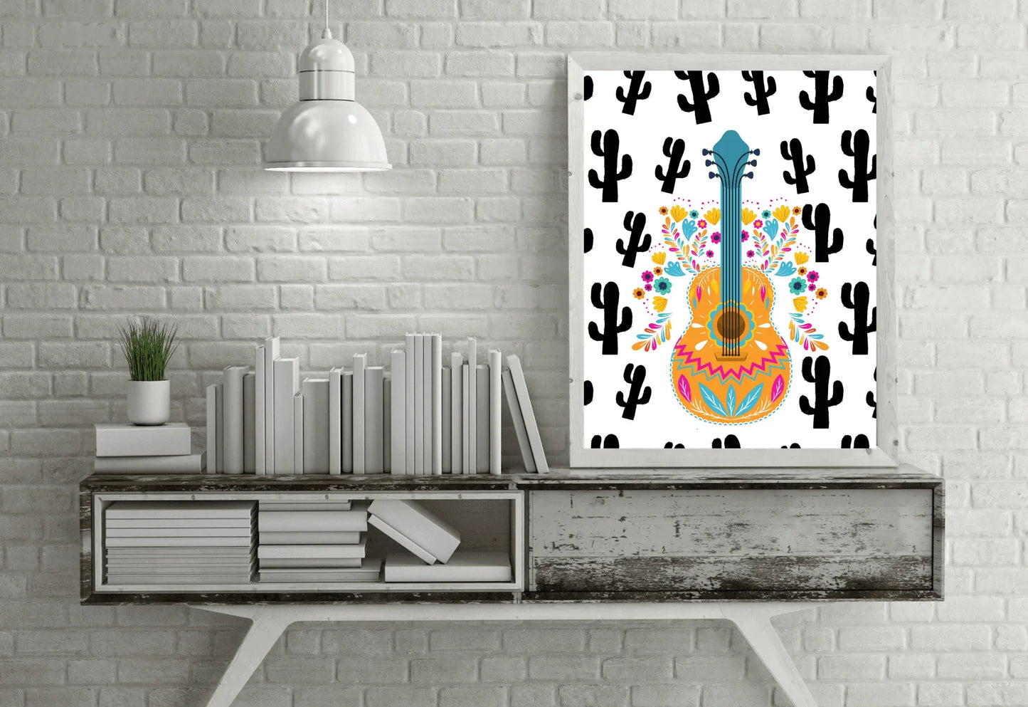 A framed boho folk art guitar print with a black cactus background, hanging on a white brick wall above a weathered wooden console table with books and a plant.
