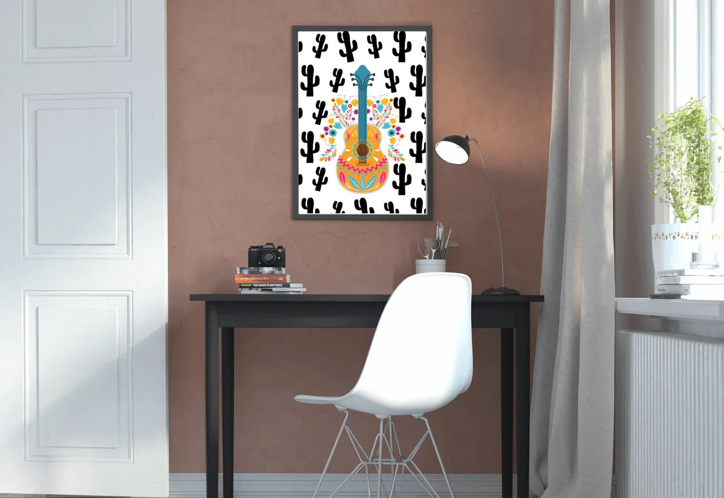 A framed boho folk art guitar print with a black cactus background, hanging on a brown wall above a black desk with a lamp and a camera.