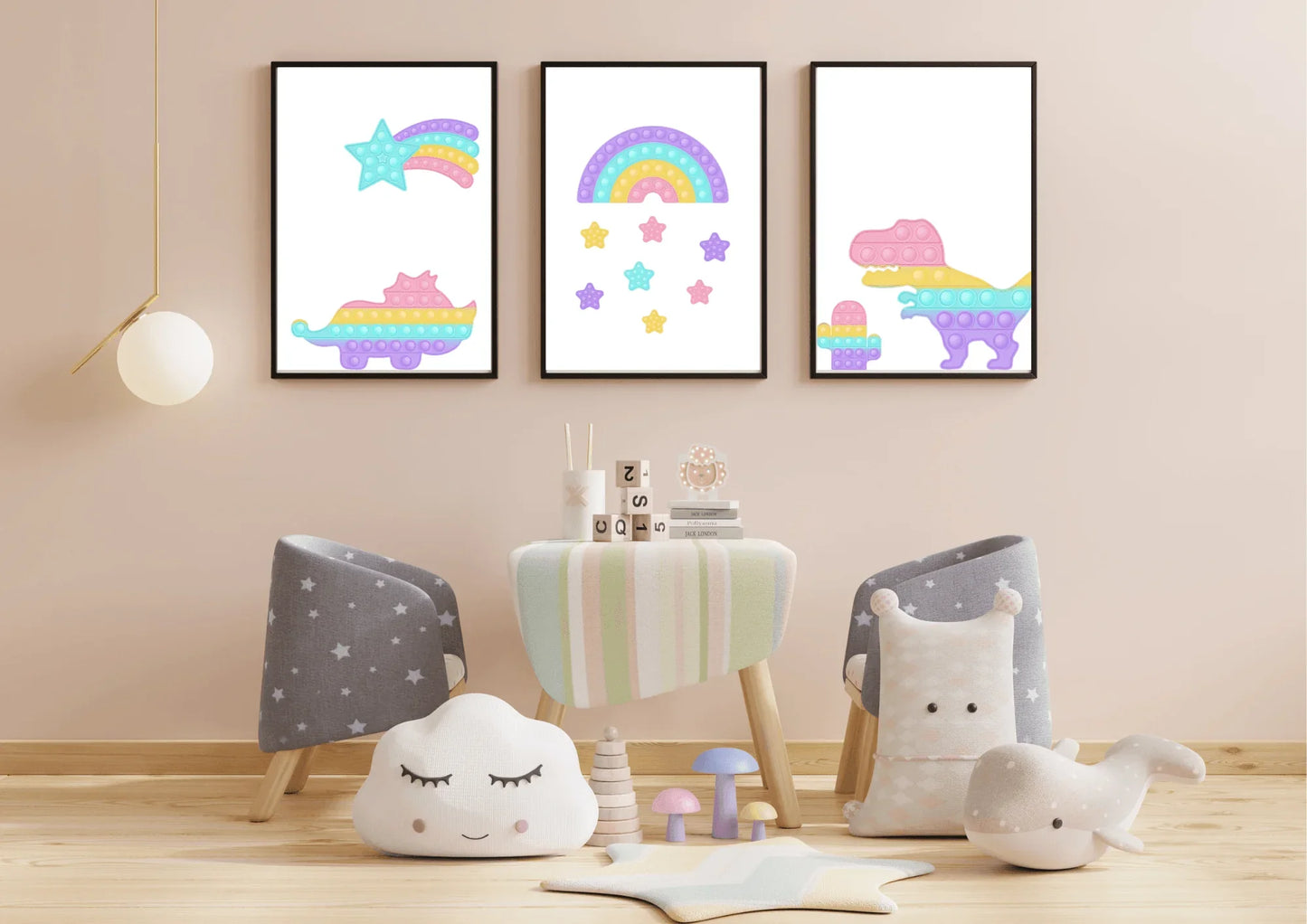 A three-piece nursery print set, featuring pop-it style dinosaurs, a rainbow, and a shooting star, hanging on a light beige wall above a small striped table with two chairs and various stuffed toys.