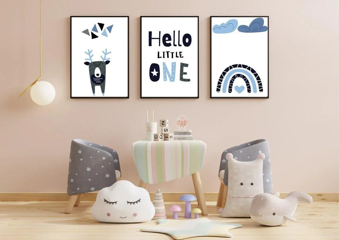A playroom with a small table and two star-patterned chairs. There are three framed prints on the wall featuring a geometric deer, the text "Hello LITTLE ONE," and a blue rainbow. Various plush toys, including a sleeping cloud and a whale, are on the floor.