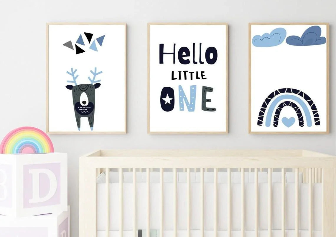 A partial view of a nursery room with a white crib. On the wall, there are three framed prints featuring a geometric deer, the text "Hello LITTLE ONE," and a blue rainbow. A colorful wooden rainbow and a white block with a letter "D" are in the foreground on the left.