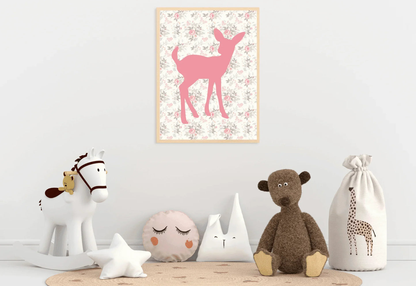A framed digital art print of a pink deer silhouette on a floral background, hanging on a white wall in a nursery. A white rocking horse, various stuffed animals and pillows, and a toy sack with a giraffe on it are arranged on the floor below the print.