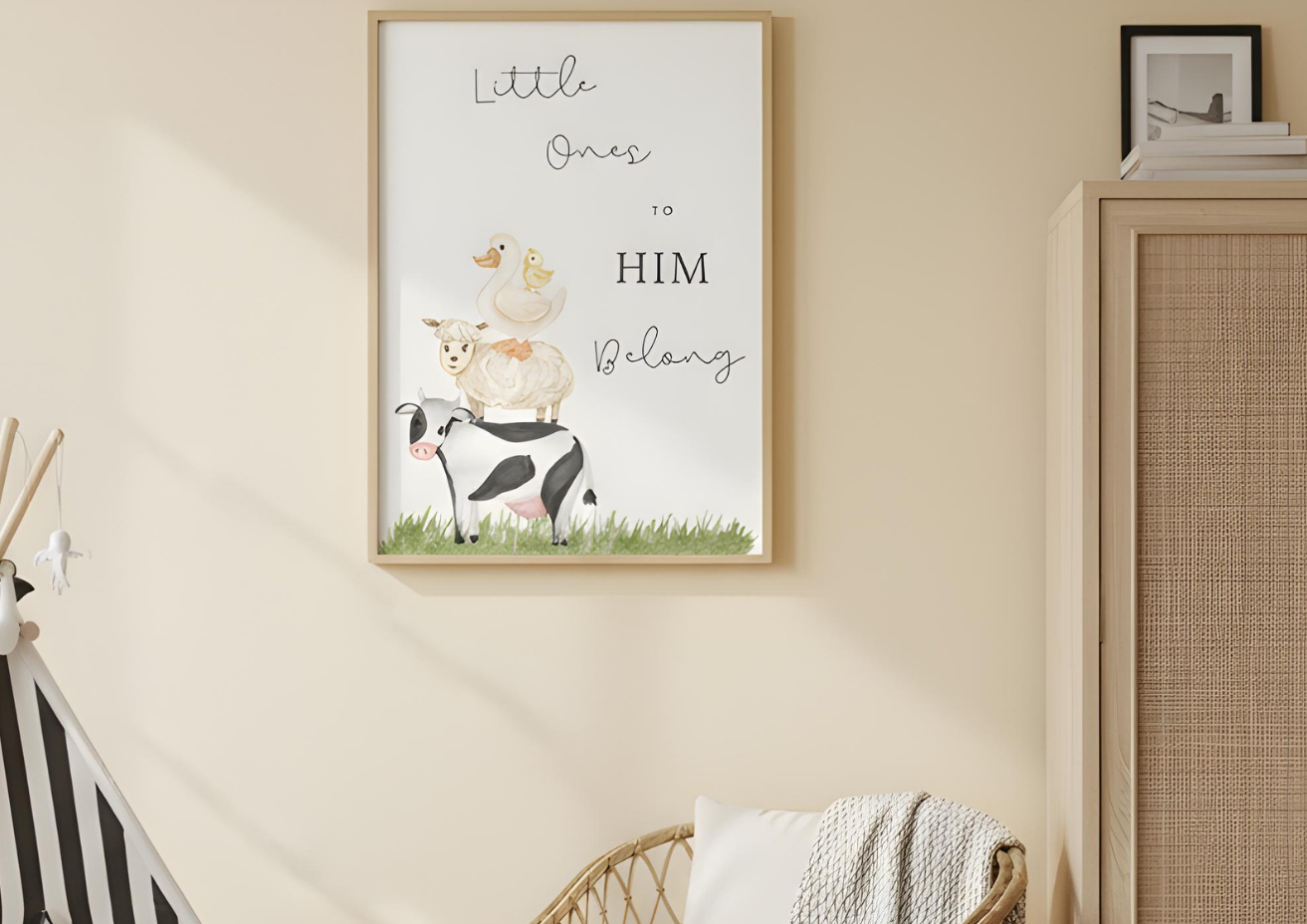A framed poster with a light wood frame hangs on a beige wall in a baby's room. The poster features a watercolor illustration of a cow, a sheep, a duck, and a chick stacked on top of each other. The text above them reads 'Little Ones to HIM Belong.'