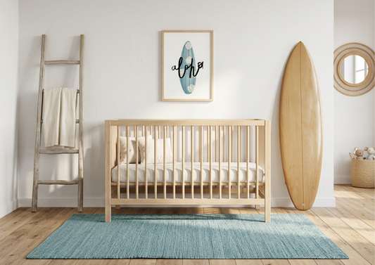 A minimalist coastal nursery featuring a light-wood spindle crib centered against a white wall. Above the crib hangs a vertical wooden-framed print of a turquoise surfboard with white monstera leaves and the word "aloha" in black script. To the right, a large natural wood surfboard leans against the wall, while a decorative wooden ladder with a cream throw sits to the left. The scene is completed with a textured turquoise-blue area rug on light wood floors, creating a bright and airy seaside aesthetic.