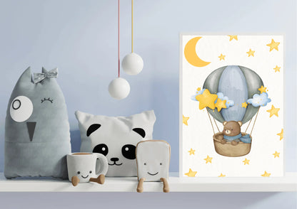 A framed vertical watercolor print displayed on a white shelf against a light blue wall, styled as part of a cozy nursery display. The artwork features a brown teddy bear wearing a blue scarf, riding in a wicker basket carried by a gray and blue hot air balloon. The white background is decorated with bright yellow stars and a crescent moon. The shelf also holds several whimsical stuffed items, including a large gray owl-shaped pillow, a panda pillow, and two small white cups or containers with smiling faces