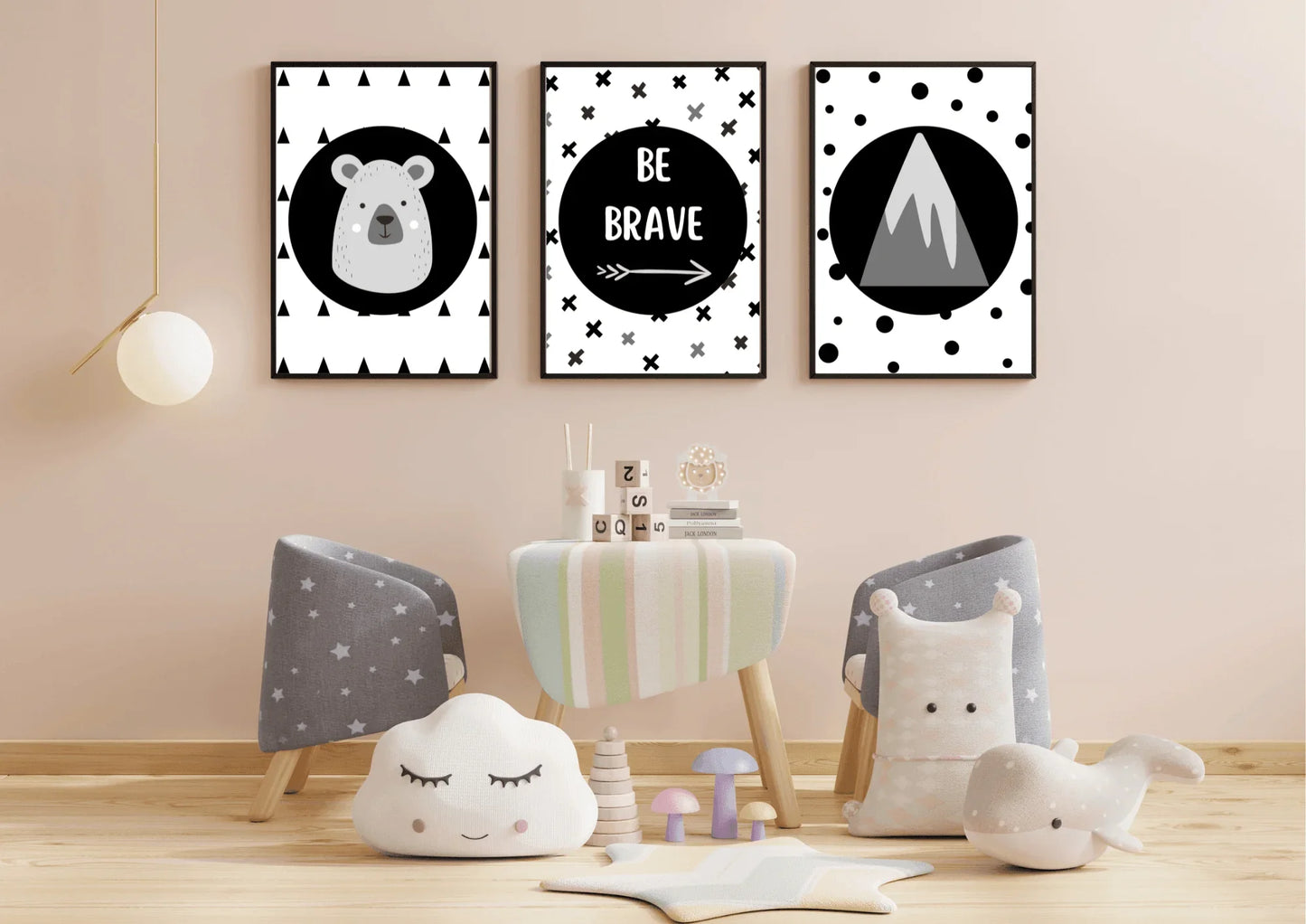 Three framed monochrome nursery prints, depicting a bear, "Be Brave" text, and a mountain, hanging on a light pink wall above a small table with children's chairs and toys.