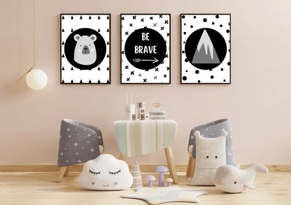 Three framed monochrome nursery prints, depicting a bear, "Be Brave" text, and a mountain, hanging on a light pink wall above a small table with children's chairs and toys.