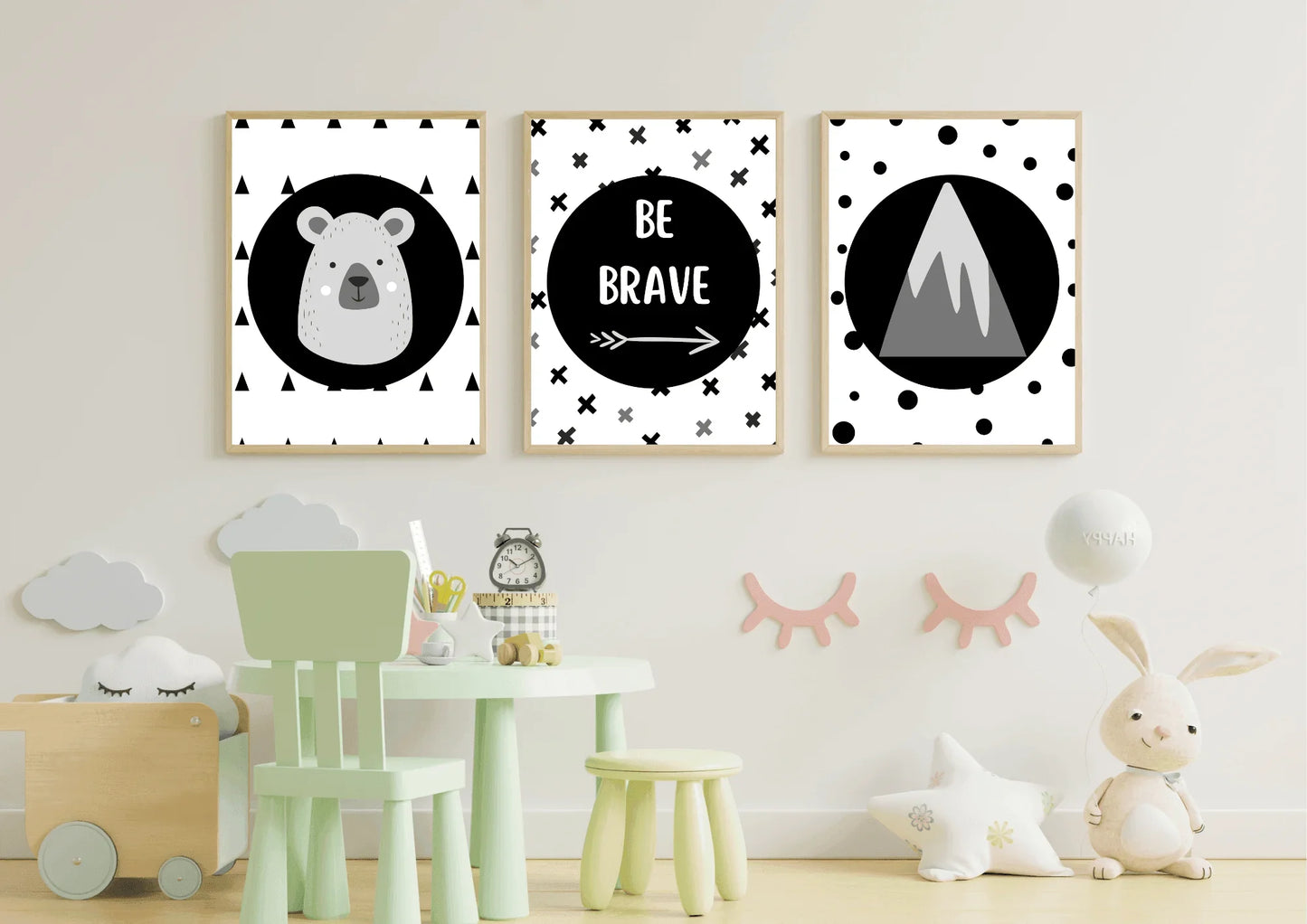 
A three-piece framed monochrome nursery print set, featuring a bear, "Be Brave" text, and a mountain, hanging above a small green table and chair set in a child's playroom.