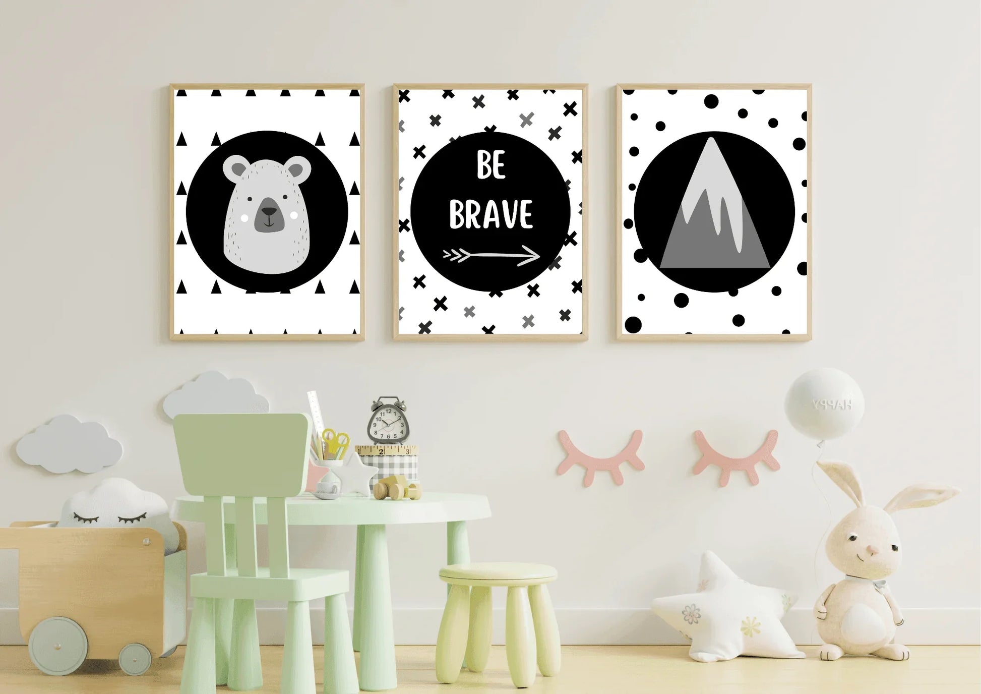 
A three-piece framed monochrome nursery print set, featuring a bear, "Be Brave" text, and a mountain, hanging above a small green table and chair set in a child's playroom.