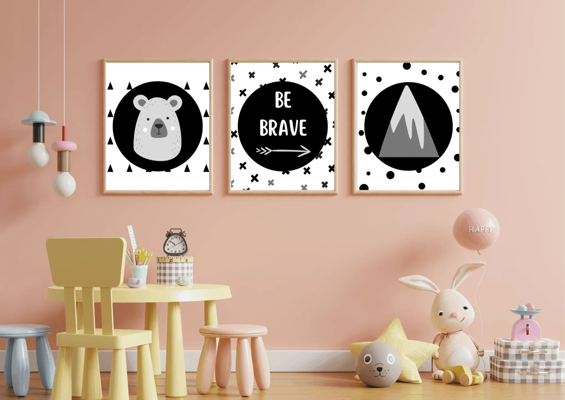 Three framed monochrome nursery prints, depicting a bear, "Be Brave" text, and a mountain, hanging on a light pink wall above a yellow table and a variety of children's toys.