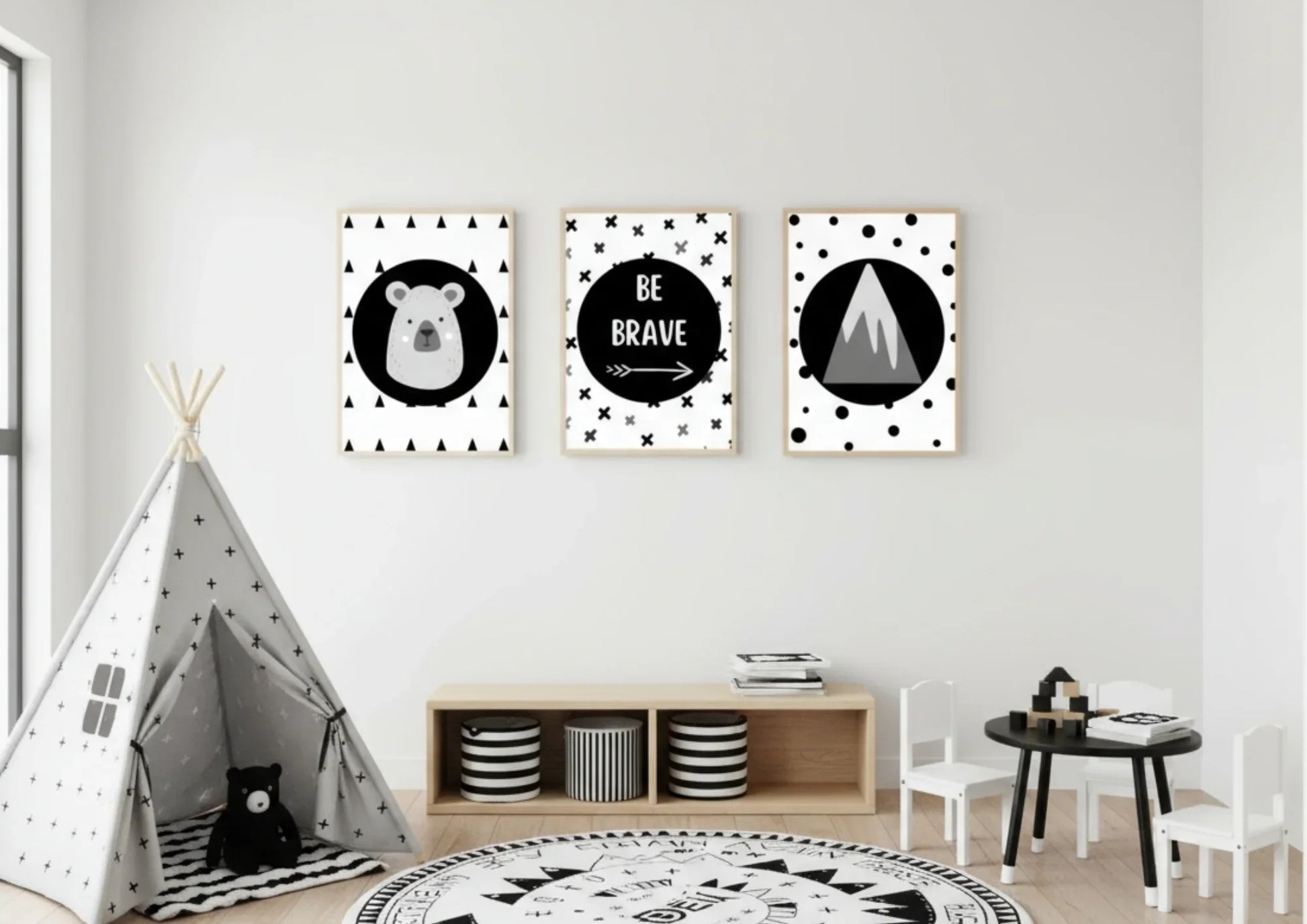 Three framed monochrome nursery prints, depicting a bear, "Be Brave" text, and a mountain, hanging on a white wall above a low shelf with a black and white striped teepee in the foreground.