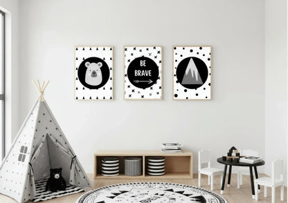 Three framed monochrome nursery prints, depicting a bear, "Be Brave" text, and a mountain, hanging on a white wall above a low shelf with a black and white striped teepee in the foreground.