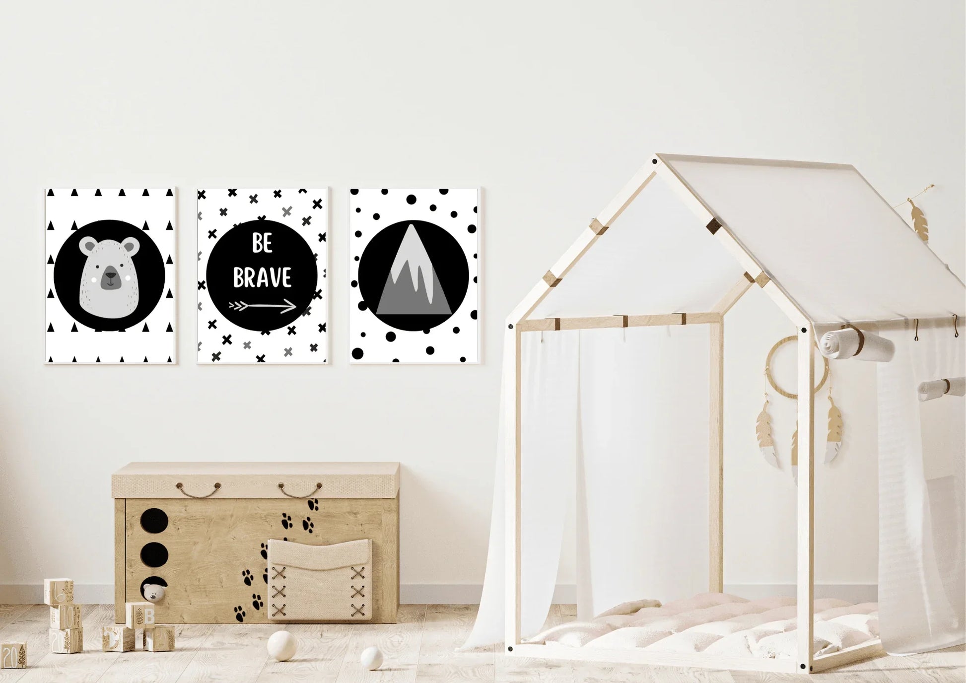Three framed monochrome nursery prints, depicting a bear, "Be Brave" text, and a mountain, hanging on a white wall above a wooden toy chest next to a bed with a canopy.