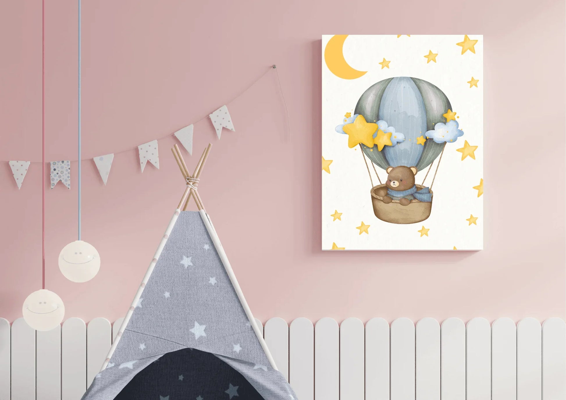 A close-up shot of a cozy nursery with pink walls and a white spindle or picket fence wainscoting. Hanging prominently on the wall is a rectangular piece of canvas art featuring a watercolor illustration of a teddy bear wearing a blue scarf, riding in a wicker basket attached to a blue-gray hot air balloon. The artwork has a light background decorated with numerous yellow stars and a large crescent moon. Below the artwork, the top of a gray and white star-patterned teepee is visible, along with a whimsical 