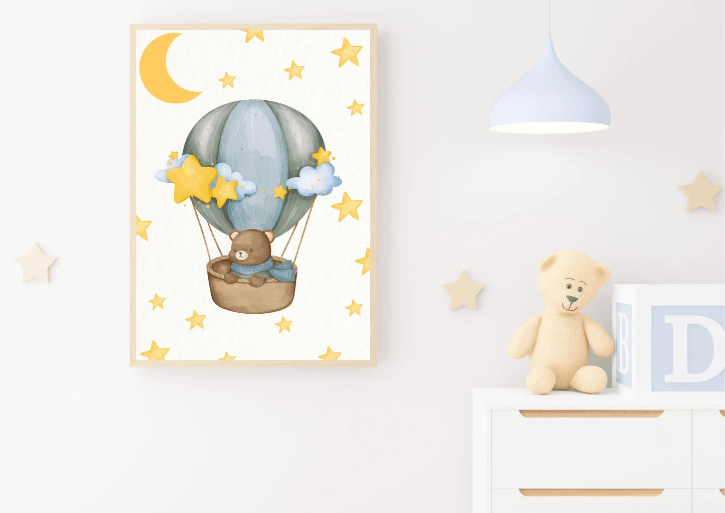 A framed vertical watercolor print displayed on a white wall in a nursery. The artwork features a cute brown teddy bear wearing a blue scarf, sitting in a wicker basket carried by a soft blue and gray hot air balloon. The background is white and patterned with yellow stars and a large yellow crescent moon. The print is styled with a simple light wood frame. To the right, on a white dresser, is a small stuffed teddy bear and large baby blocks. A light blue, modern pendant lamp hangs from the ceiling.