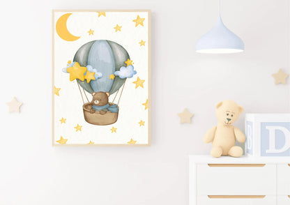 A framed vertical watercolor print displayed on a white wall in a nursery. The artwork features a cute brown teddy bear wearing a blue scarf, sitting in a wicker basket carried by a soft blue and gray hot air balloon. The background is white and patterned with yellow stars and a large yellow crescent moon. The print is styled with a simple light wood frame. To the right, on a white dresser, is a small stuffed teddy bear and large baby blocks. A light blue, modern pendant lamp hangs from the ceiling.