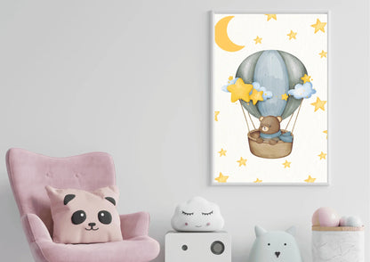 A vertical framed watercolor print hanging on a white wall in a child's room. The artwork features a sweet brown teddy bear wearing a blue scarf, sitting comfortably in a wicker basket carried by a soft gray and blue hot air balloon. The light-colored background is filled with scattered yellow stars and a large crescent moon at the top. Below the artwork is a small white cube organizer holding cute, plush toys, including a cloud and an animal. To the left, a portion of a plush pink armchair with a panda-the