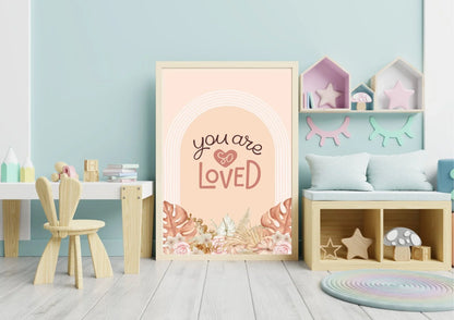 
A wooden-framed "You are so LOVED" boho art print on the floor of a playroom with a small desk and a storage bench.