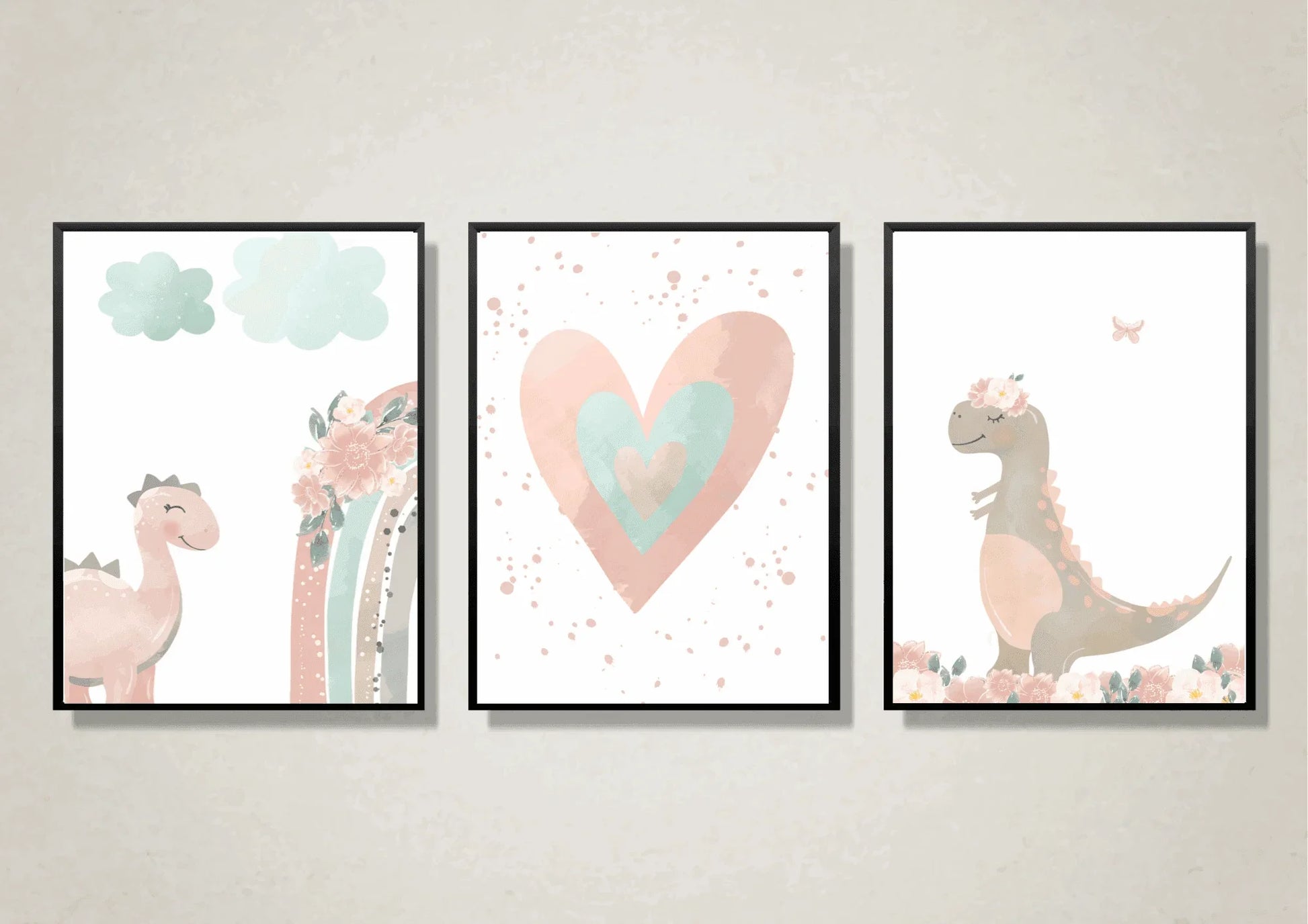 A close-up view of three framed art prints hanging on a light, textured wall. The prints are a whimsical, watercolor set featuring a pink dinosaur, a pink and teal heart, and a gray dinosaur with a floral crown. Each is framed in a thin black frame.