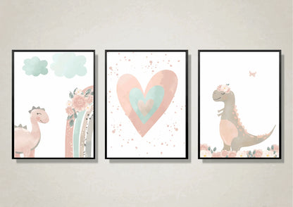 A close-up view of three framed art prints hanging on a light, textured wall. The prints are a whimsical, watercolor set featuring a pink dinosaur, a pink and teal heart, and a gray dinosaur with a floral crown. Each is framed in a thin black frame.