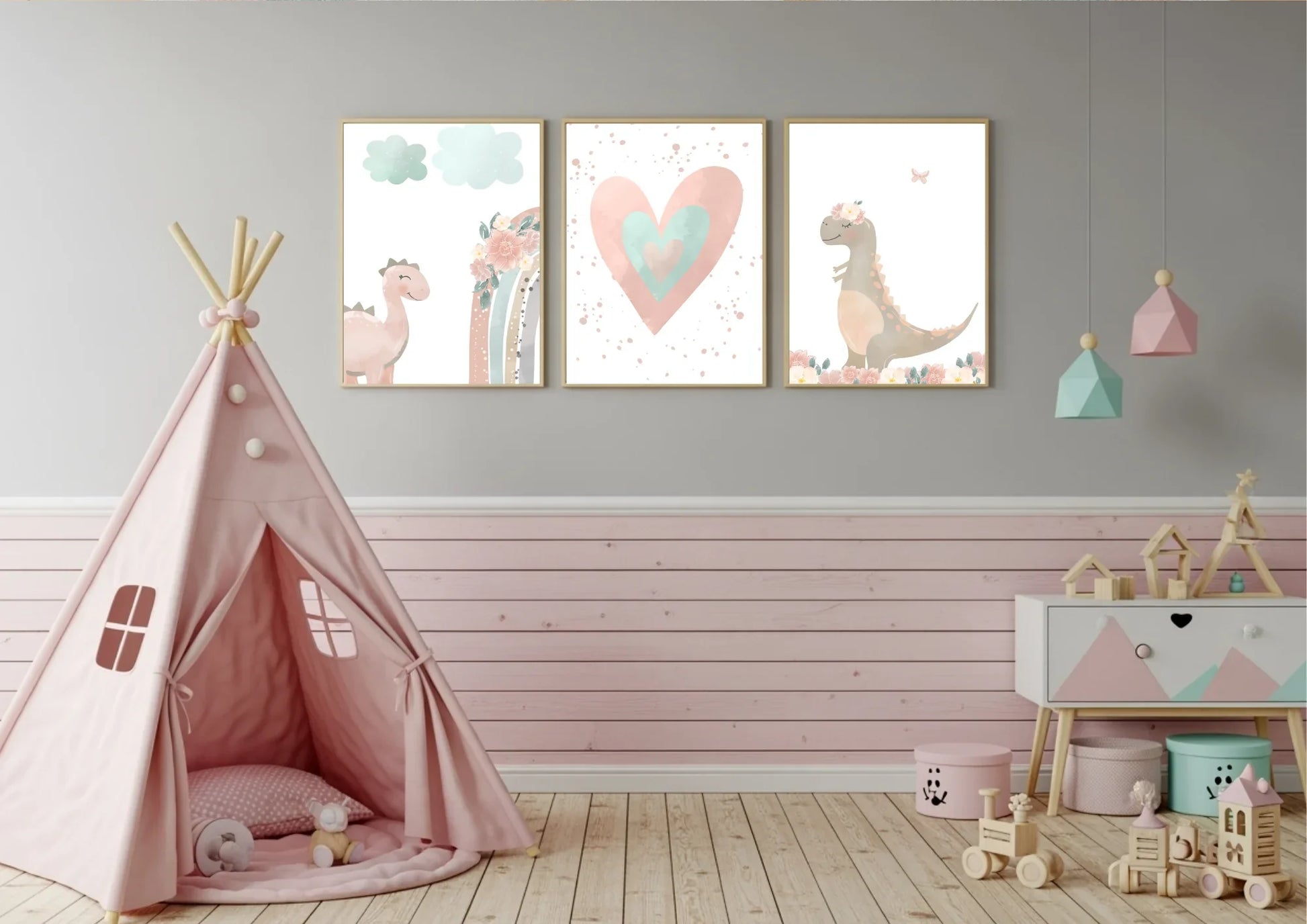 A set of three framed nursery prints featuring a pink dinosaur, a rainbow, a large heart, and a dinosaur with a floral crown, hanging on a gray and pink slatted wall. A pink teepee tent is on the left, and a white and pink dresser with various toys is on the right.