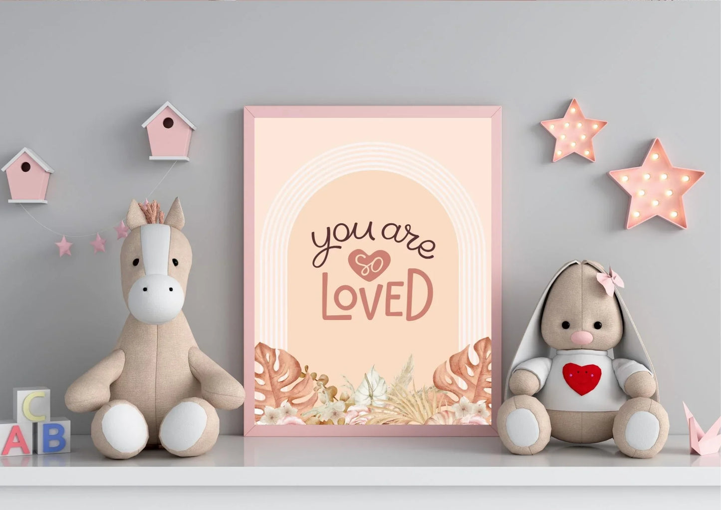 
A framed "You are so LOVED" boho art print on a shelf with stuffed animals and pink star lights.